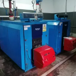 Dwa niebieskie kotły gazowe Buderus Logano GE515 z czerwonymi palnikami, widoczne w kotłowni z szaro-białymi ścianami i płytkami podłogowymi, z tabliczką ostrzegawczą o napięciu elektrycznym.