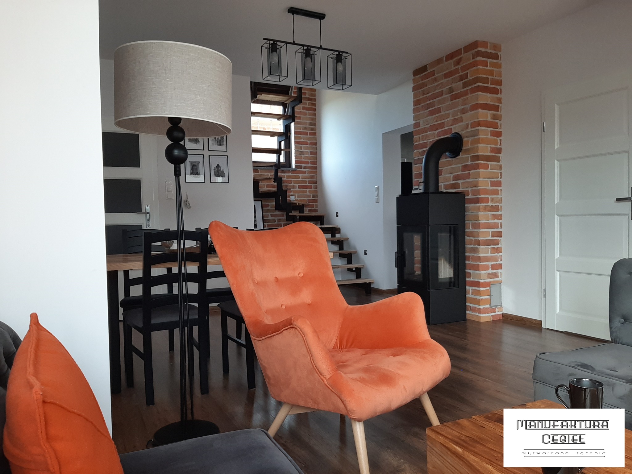 Wnętrze salonu z pomarańczowym fotelem na drewnianych nogach, czarnym kominkiem obudowanym cegłą, drewnianymi schodami i industrialną lampą wiszącą.