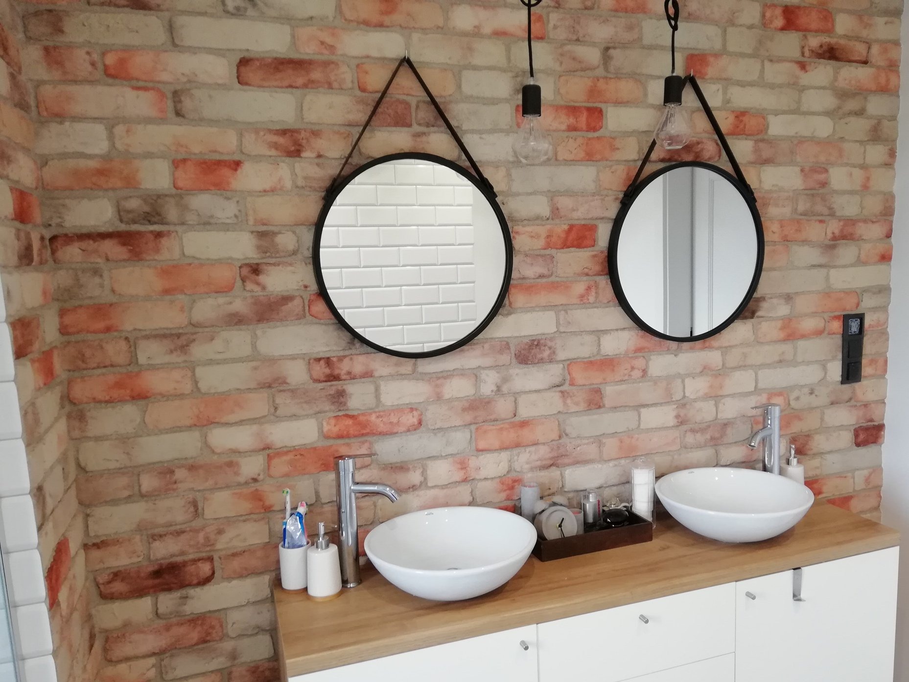 Nowoczesna łazienka z dwoma okrągłymi lustrami zawieszonymi na paskach, umywalki nablatowe na drewnianym blacie i ścianą z cegły w odcieniach czerwieni i bieli.