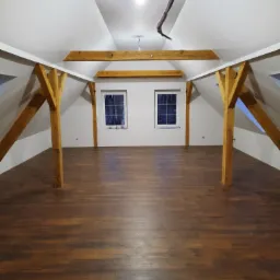 Poddasze po remoncie z drewnianą podłogą, bielonymi ścianami i odsłoniętymi drewnianymi belkami konstrukcyjnymi, widok na dwa okna w białych ramach.