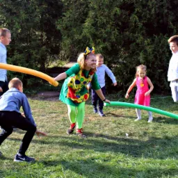 Kobieta w stroju klauna z kolorowymi łatami na fartuchu i zielonych rajstopach, bawi się z grupą dzieci na trawie, używając długich, piankowych rurek w kolorach żółtym i zielonym.