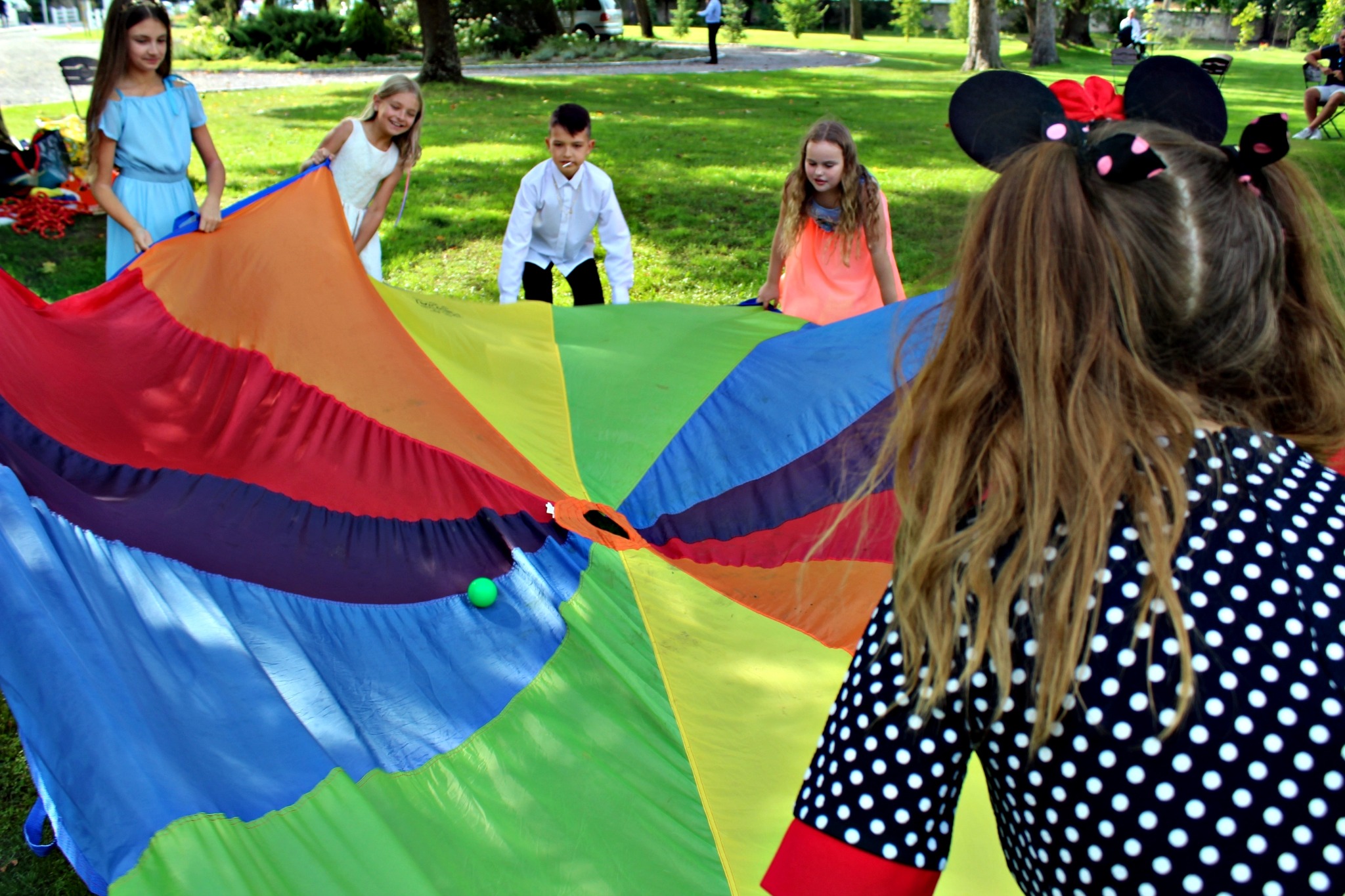 Dzieci trzymające kolorową chustę animacyjną na trawie, widok zza dziecka w bluzce w grochy i uszach Myszki Miki.