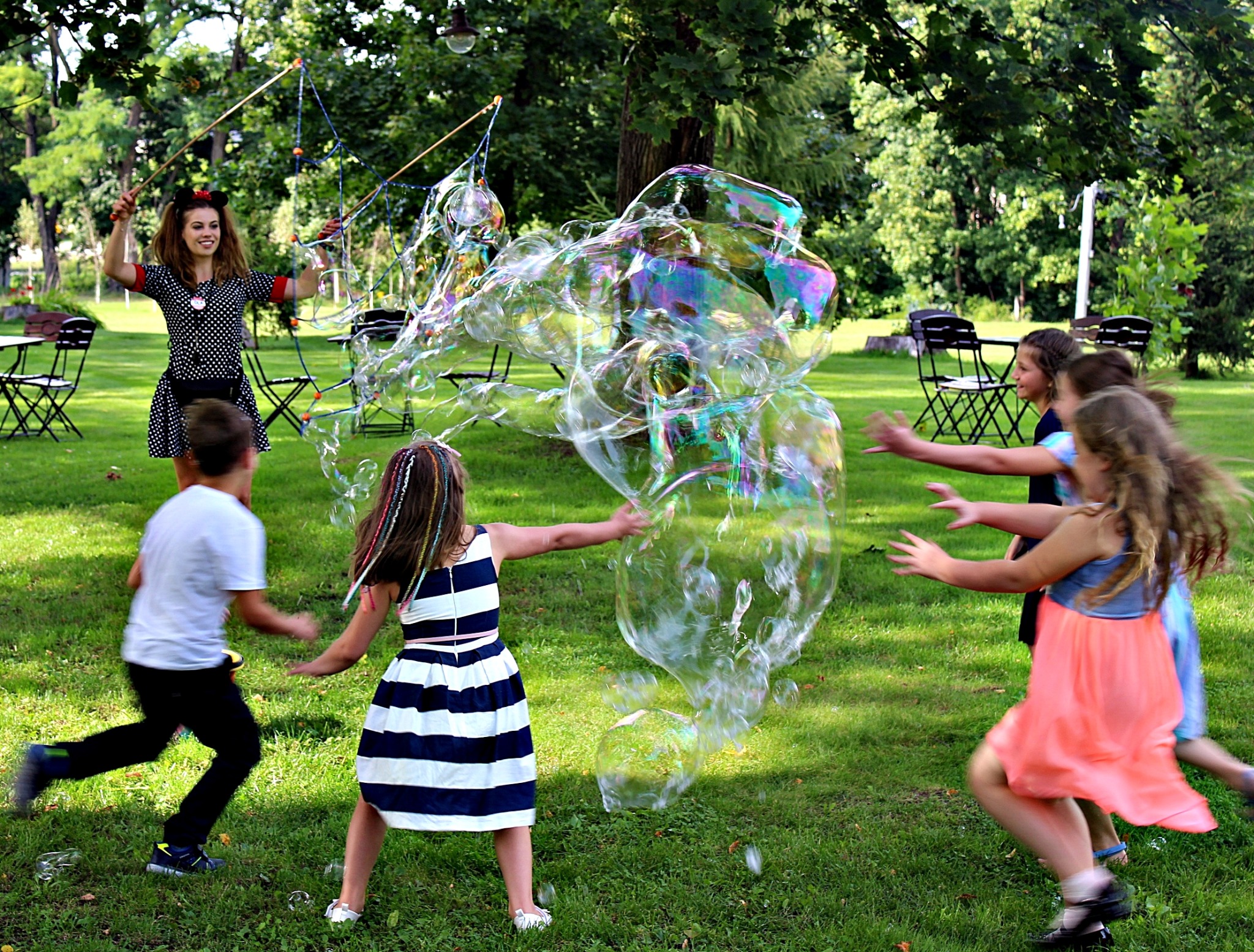 Kobieta w stroju w kropki tworzy gigantyczne bańki mydlane na trawie, wokół biegają dzieci próbujące je złapać, w tle stoliki piknikowe.