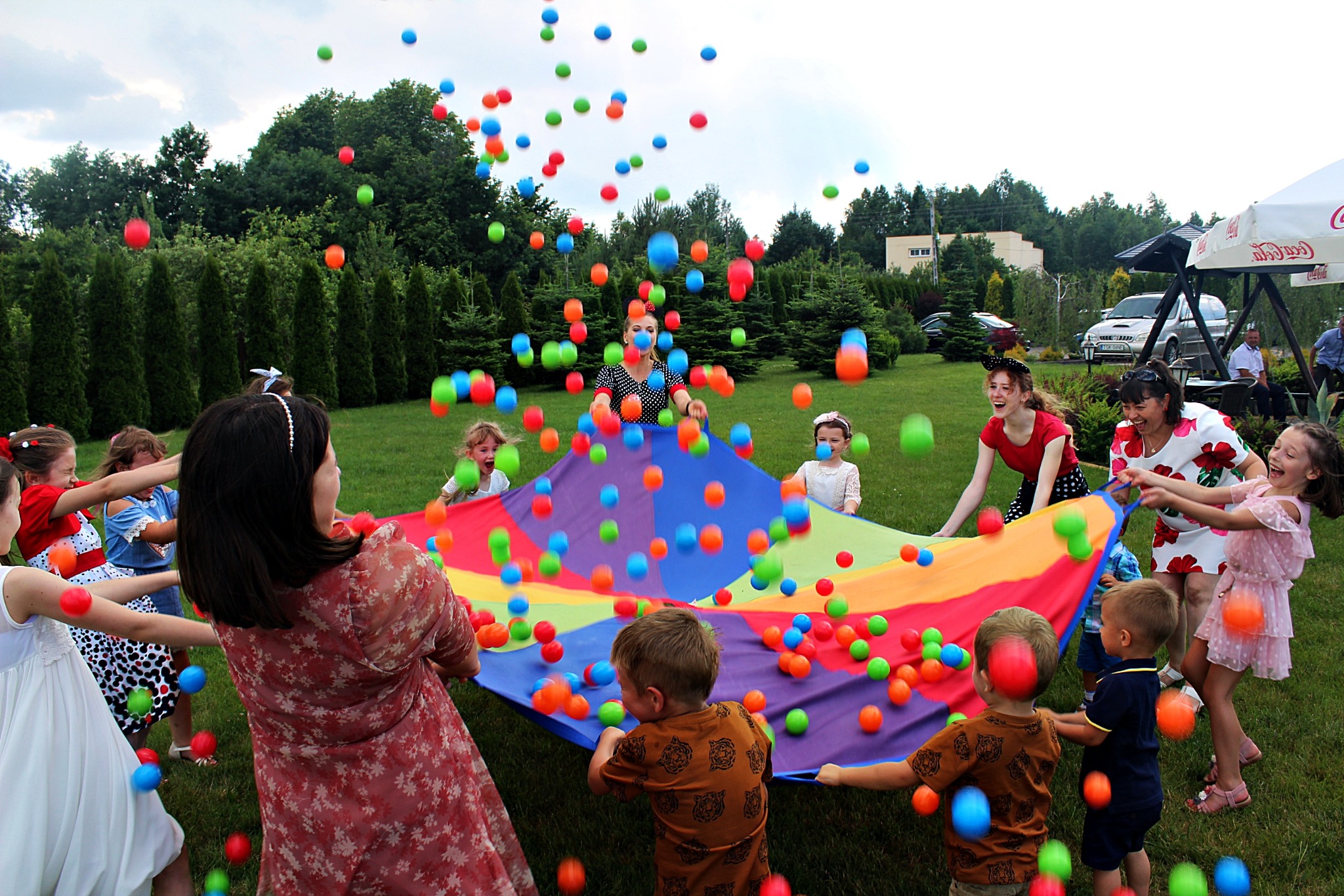 Grupa dzieci i dorosłych bawi się na trawie kolorową chustą animacyjną, podrzucając w górę mnóstwo kolorowych piłeczek.