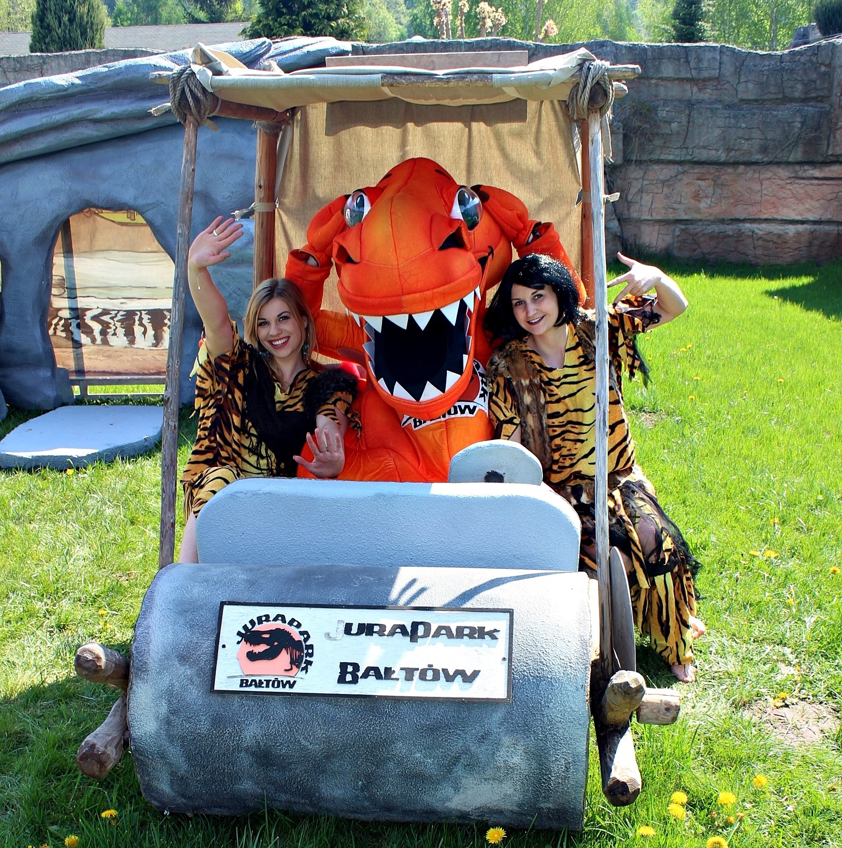 Animatorzy w strojach jaskiniowców i maskotka dinozaura w wozie z kamienia na tle Juraparku Bałtów