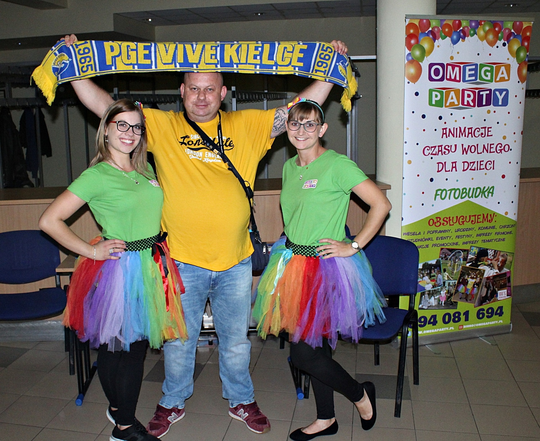 Ekipa eventowa w kolorowych tutu i koszulkach z logo firmy, mężczyzna trzyma szalik kibica PGE Vive Kielce, w tle baner reklamowy z napisem 'Animacje czasu wolnego dla dzieci, Fotobudka'.