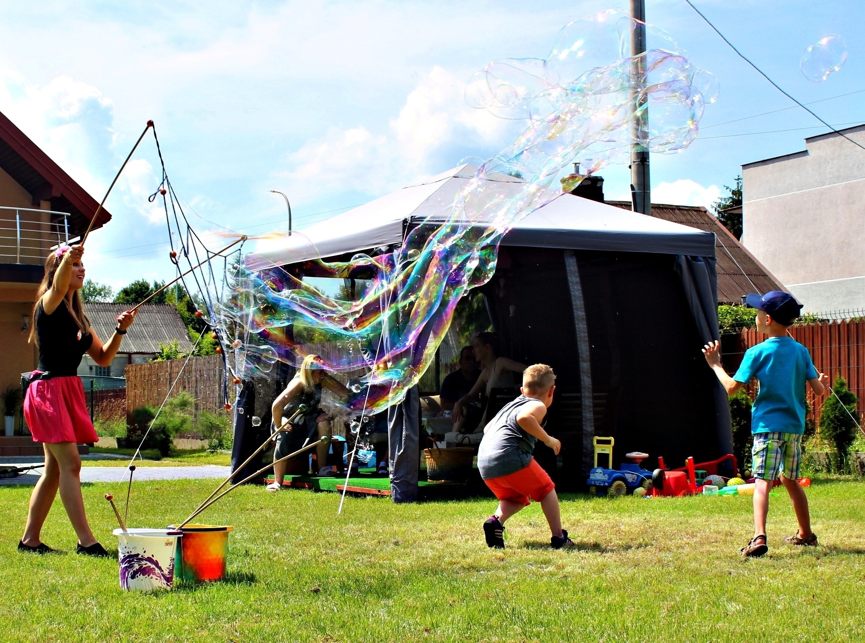 Kobieta tworzy gigantyczne bańki mydlane na zielonej trawie, dzieci obserwują, w tle namiot i dom.