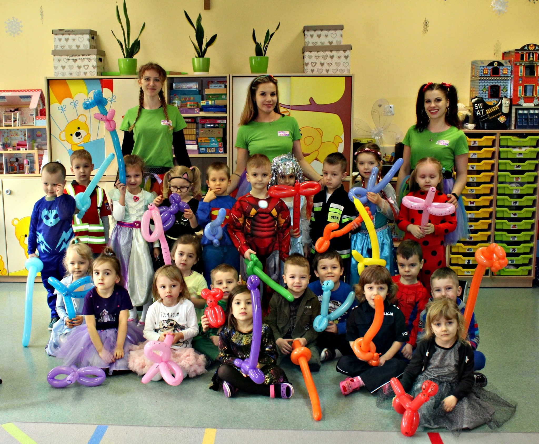 Grupa dzieci w kostiumach i z balonami modelowanymi w kształty zwierząt i przedmiotów, pozujących z trzema kobietami w zielonych koszulkach z logo firmy, na tle sali zabaw z regałami i zabawkami.