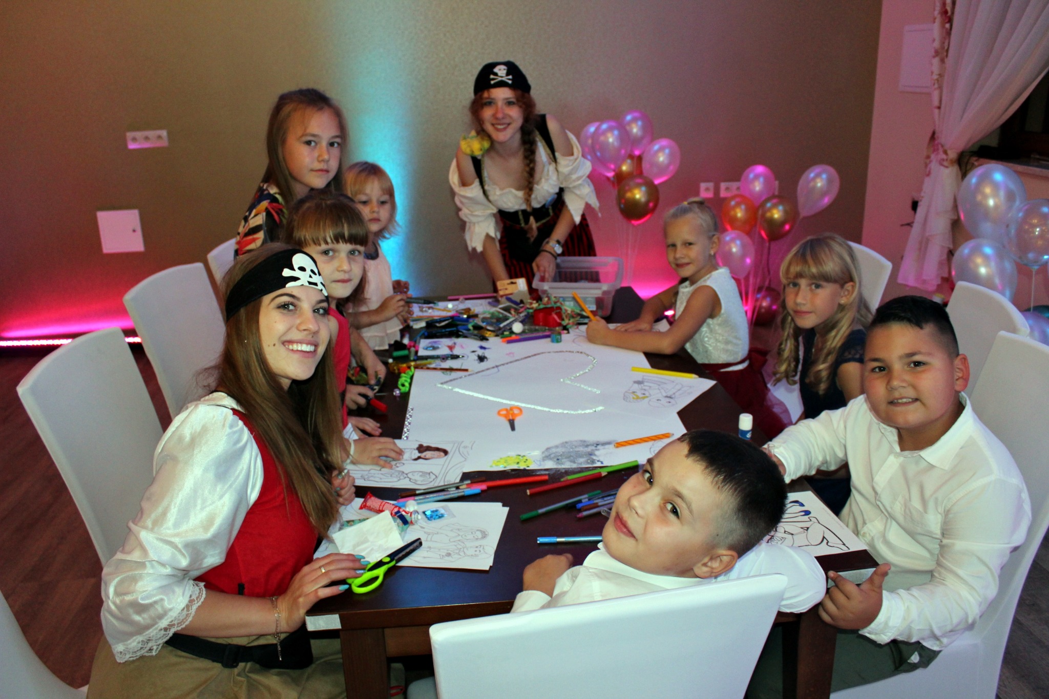 Grupa dzieci i animatorki w strojach pirackich rysują i kolorują przy stole podczas imprezy urodzinowej, w tle balony.
