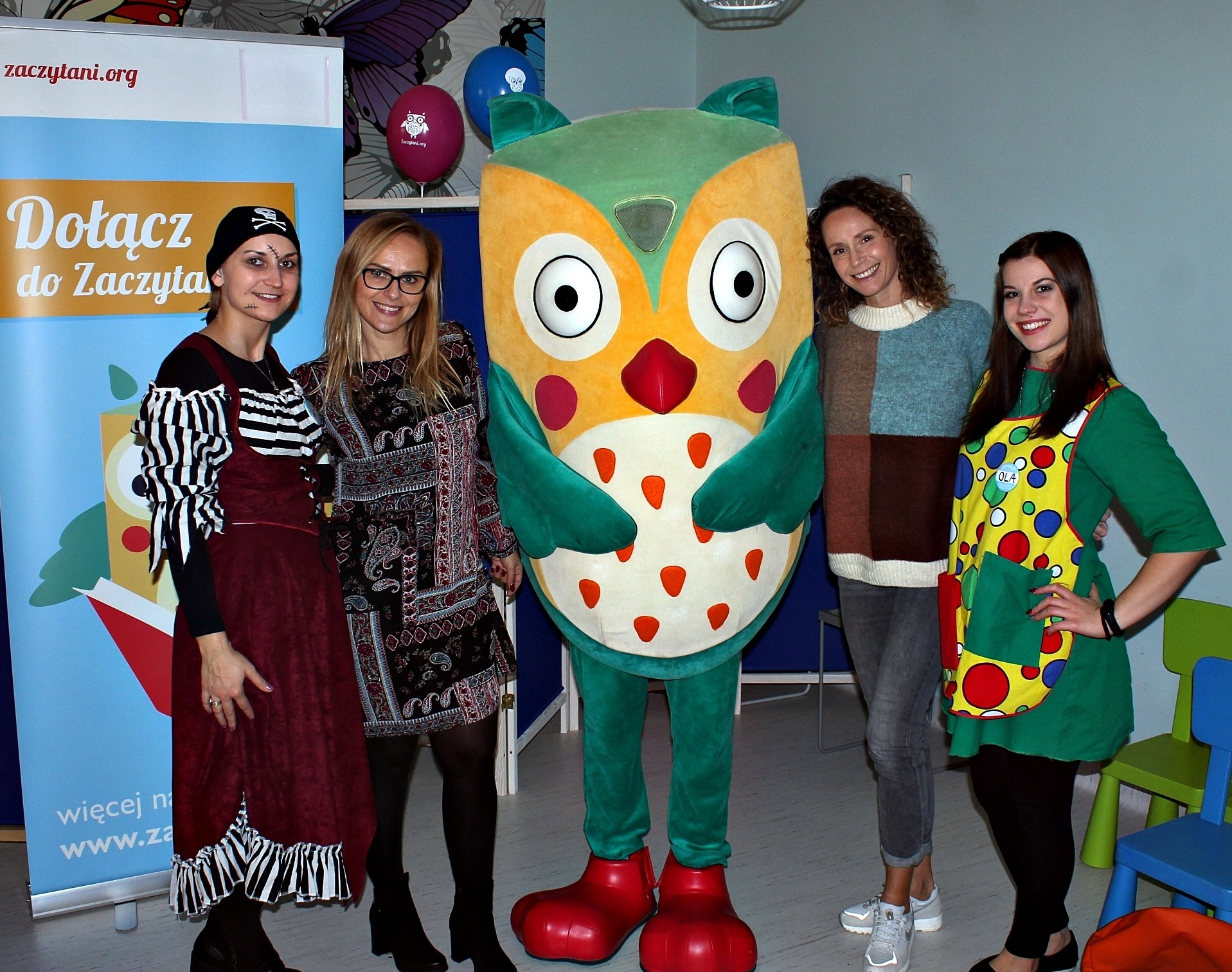 Grupa czterech osób, w tym jedna w stroju pluszowej sowy, pozuje na tle plakatu i balonów z logo 'Zaczytani.org', kobieta po lewej w kostiumie pirata z pomalowaną blizną na twarzy.