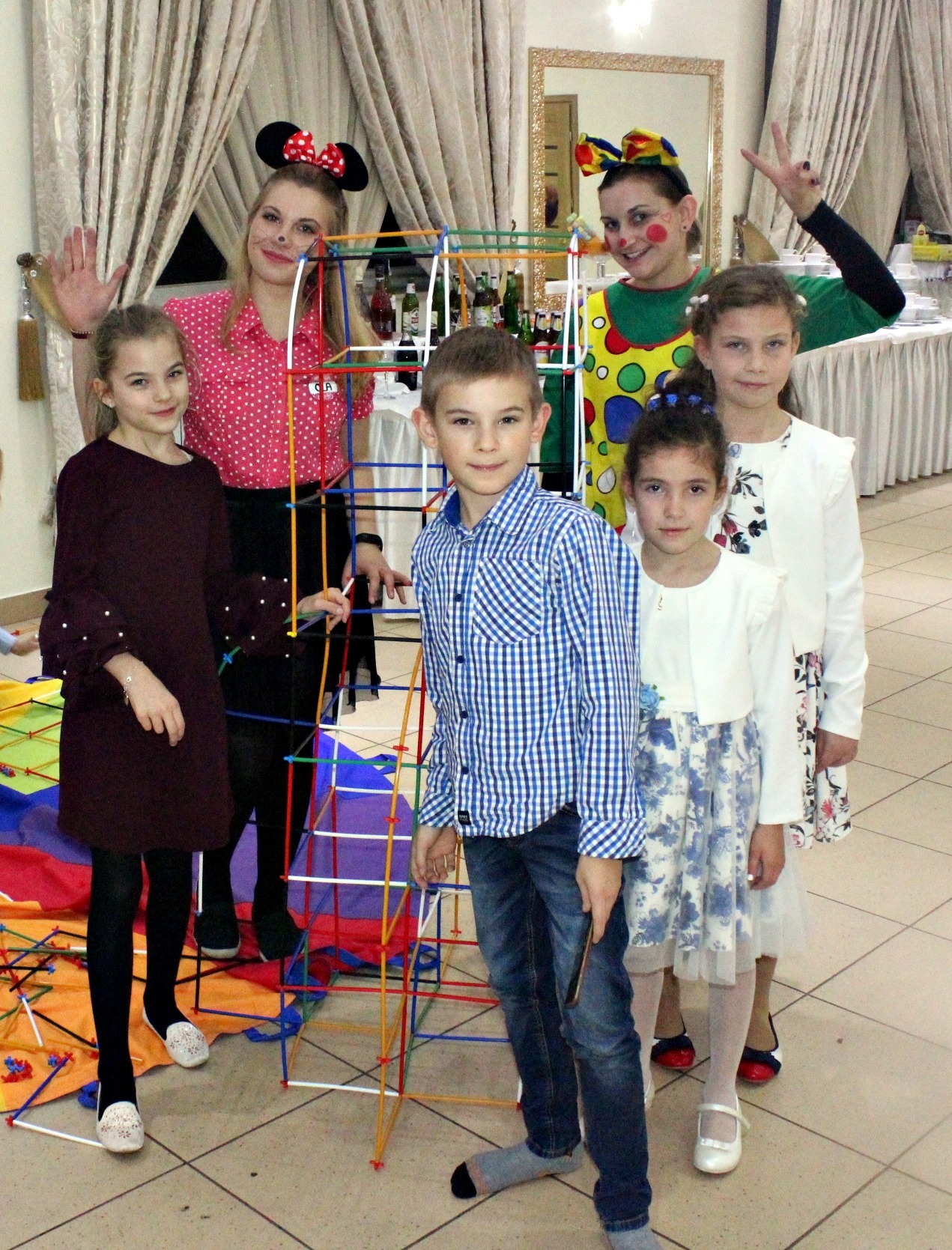 Grupa dzieci z animatorkami w kostiumach Myszki Miki i klauna pozuje przy kolorowej konstrukcji z rurek w pomieszczeniu z zasłonami i stołem z przekąskami w tle.