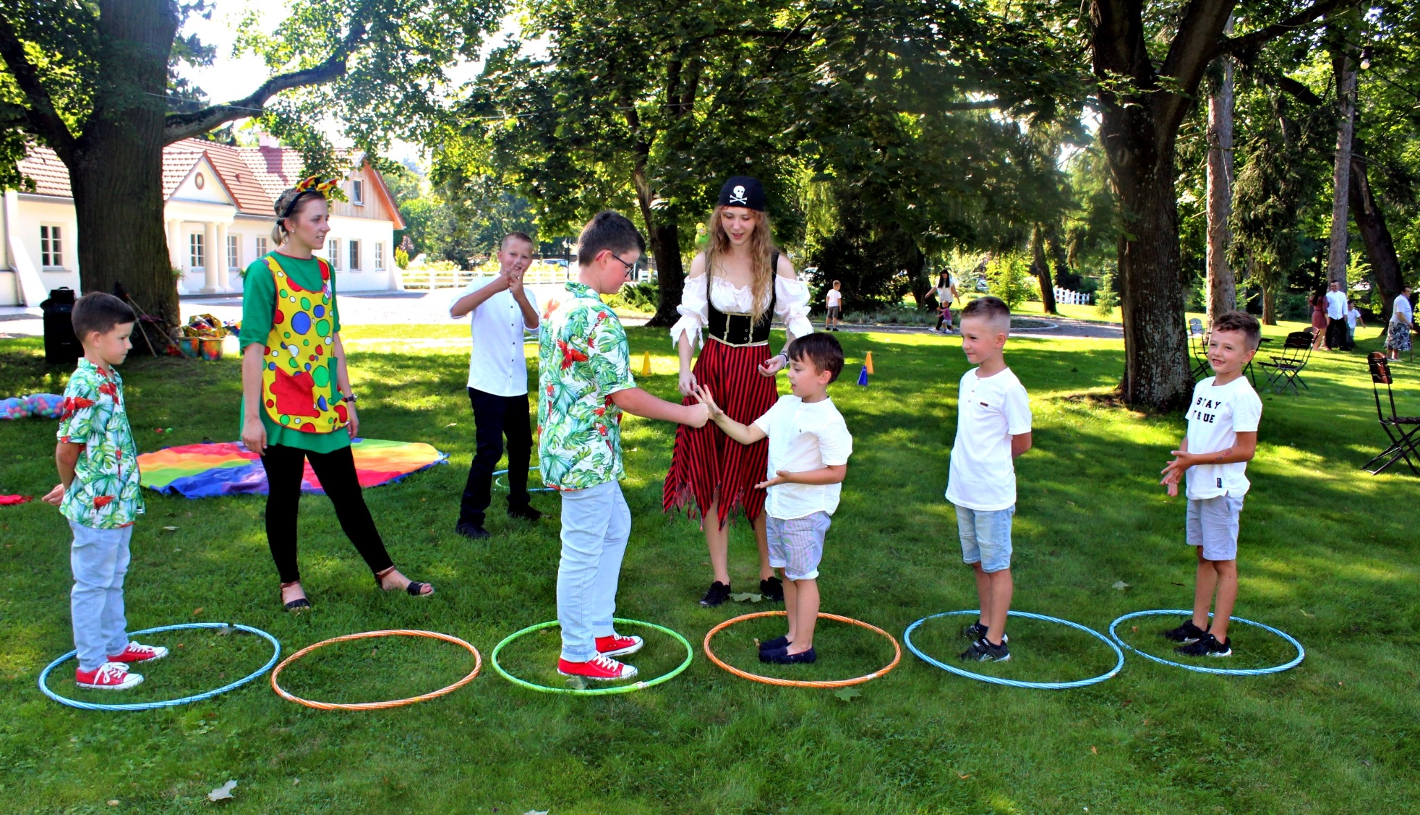 Animatorzy w strojach pirata i klauna prowadzą gry z dziećmi na trawie, wykorzystując kolorowe hula-hoop, w tle budynek z czerwonym dachem.
