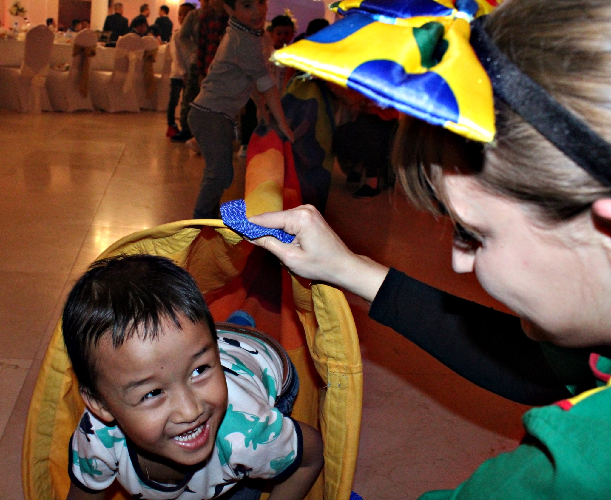 Uśmiechnięty chłopiec w tunelu z kolorowej tkaniny, trzymanym przez osobę w kostiumie z żółto-niebieską ozdobą na głowie, zabawa na imprezie.