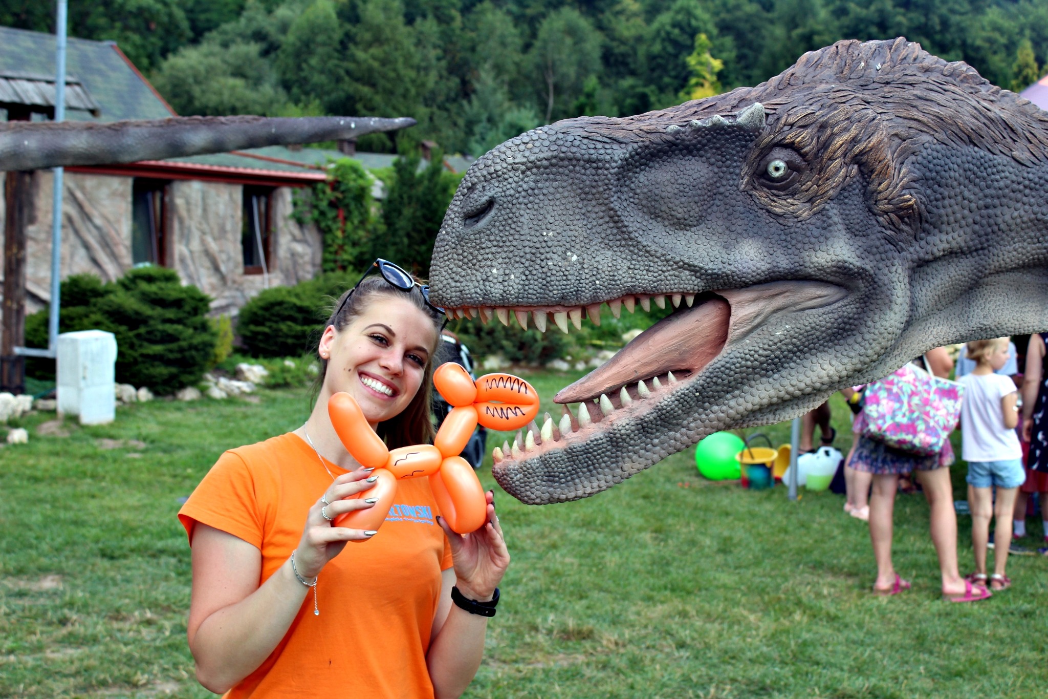 Uśmiechnięta kobieta w pomarańczowej koszulce trzyma balonowego pieska przed otwartą paszczą modelu dinozaura w parku rozrywki.