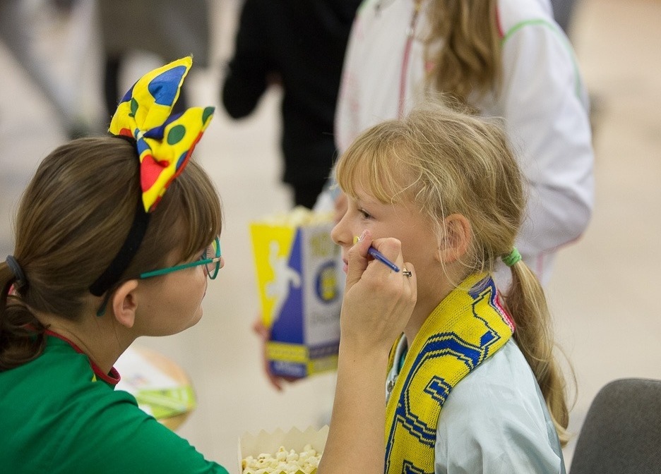 Dziewczynka z blond włosami, w trakcie malowania twarzy, z żółto-niebieskim szalikiem na szyi, w tle osoba z popcornem i dziewczynka w zielonej bluzce z kolorową opaską na głowie.