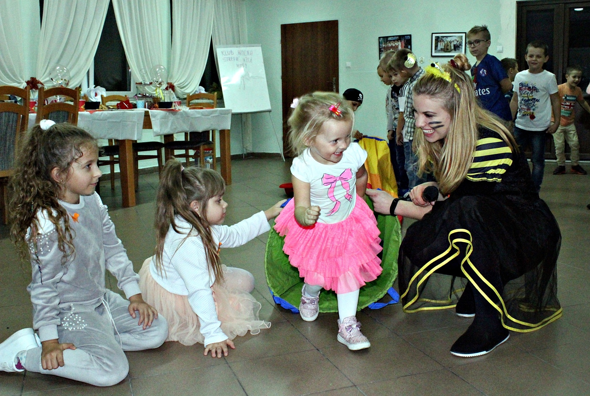 Dziewczynka w różowej tutu spódniczce przechodzi przez tunel animacyjny, wspierana przez animatorkę w czarno-żółtym stroju pszczoły, podczas zabawy z innymi dziećmi na tle sali z dekoracjami.