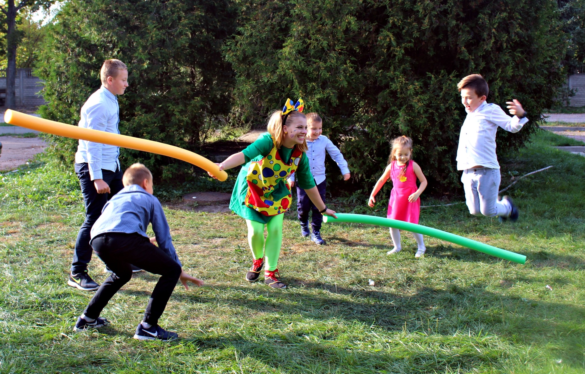 Kobieta w stroju klauna z kolorowymi łatami na fartuchu i zielonych rajstopach, bawi się z grupą dzieci na trawie, używając długich, piankowych rurek w kolorach żółtym i zielonym.
