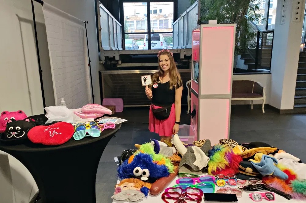 Młoda kobieta w różowej spódnicy trzymająca rekwizyt z parą młodą przed różową fotobudką, na stole leżą kolorowe rekwizyty do zdjęć, w tle industrialne wnętrze.