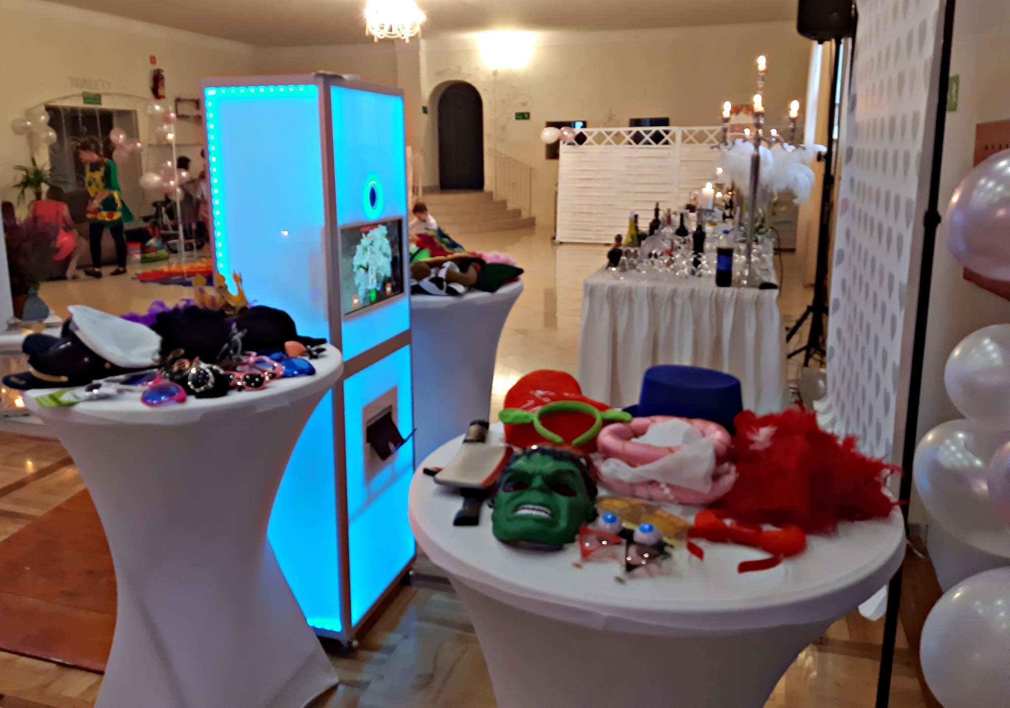 Przygotowane rekwizyty do fotobudki: maska Hulka, kolorowe okulary, kapelusze i boa na białych stolikach z podświetlaną fotobudką w tle, widoczny fragment dekoracji sali z balonami i stołem...