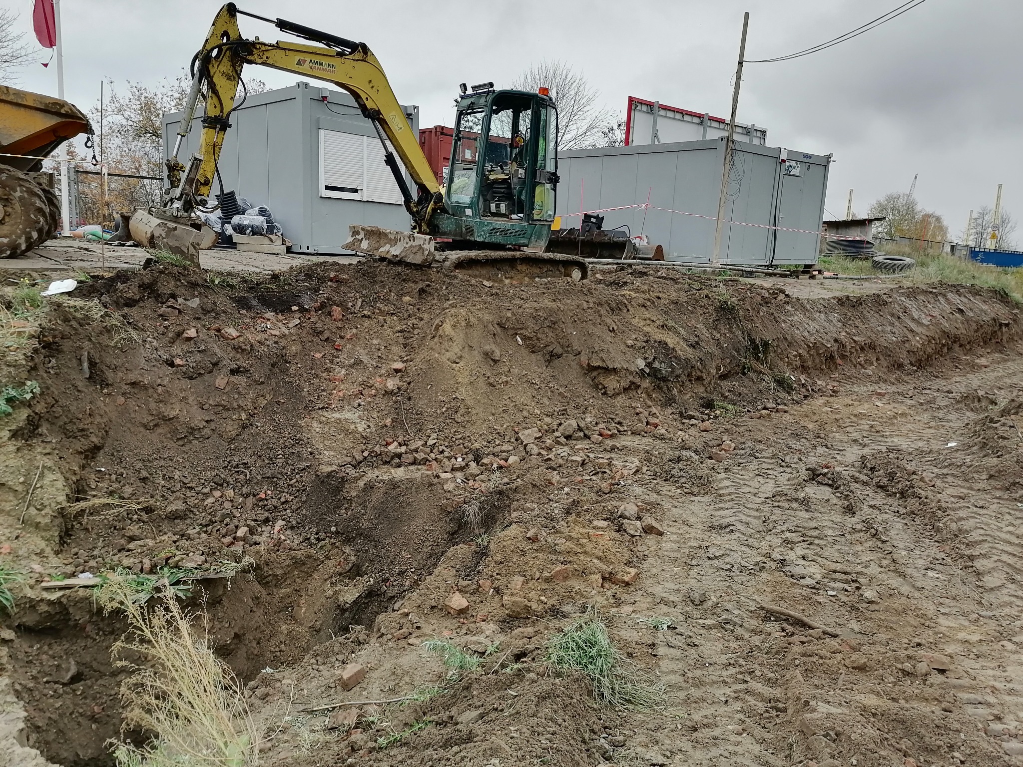Mała koparka gąsienicowa na placu budowy, tuż obok wykopu pod instalacje, z kontenerami biurowymi w tle i widocznymi śladami opon na ziemi.