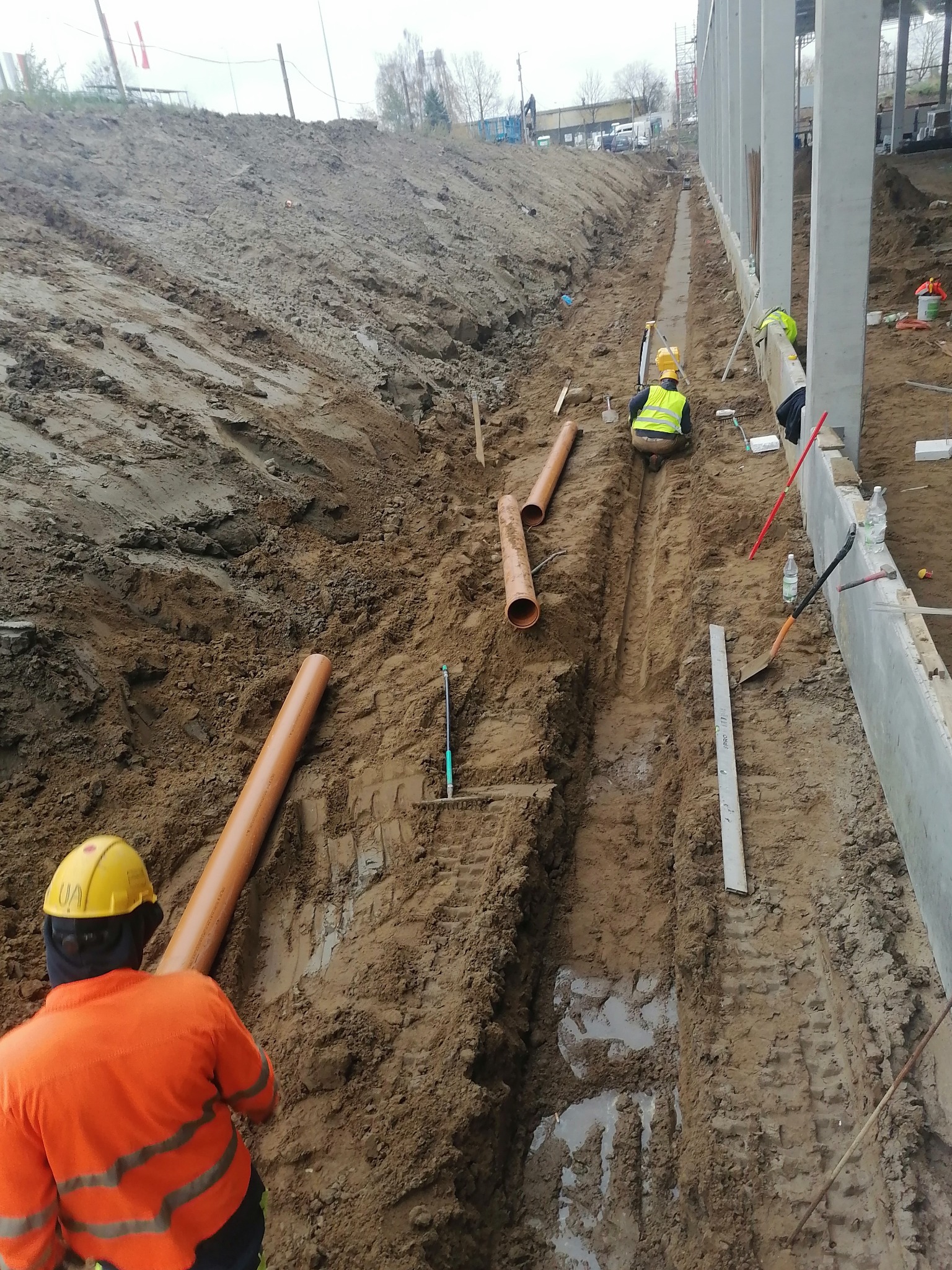 Pracownicy w odblaskowych kamizelkach układają pomarańczowe rury w wykopie wzdłuż betonowej konstrukcji, widoczne narzędzia i ślady opon na mokrej ziemi.