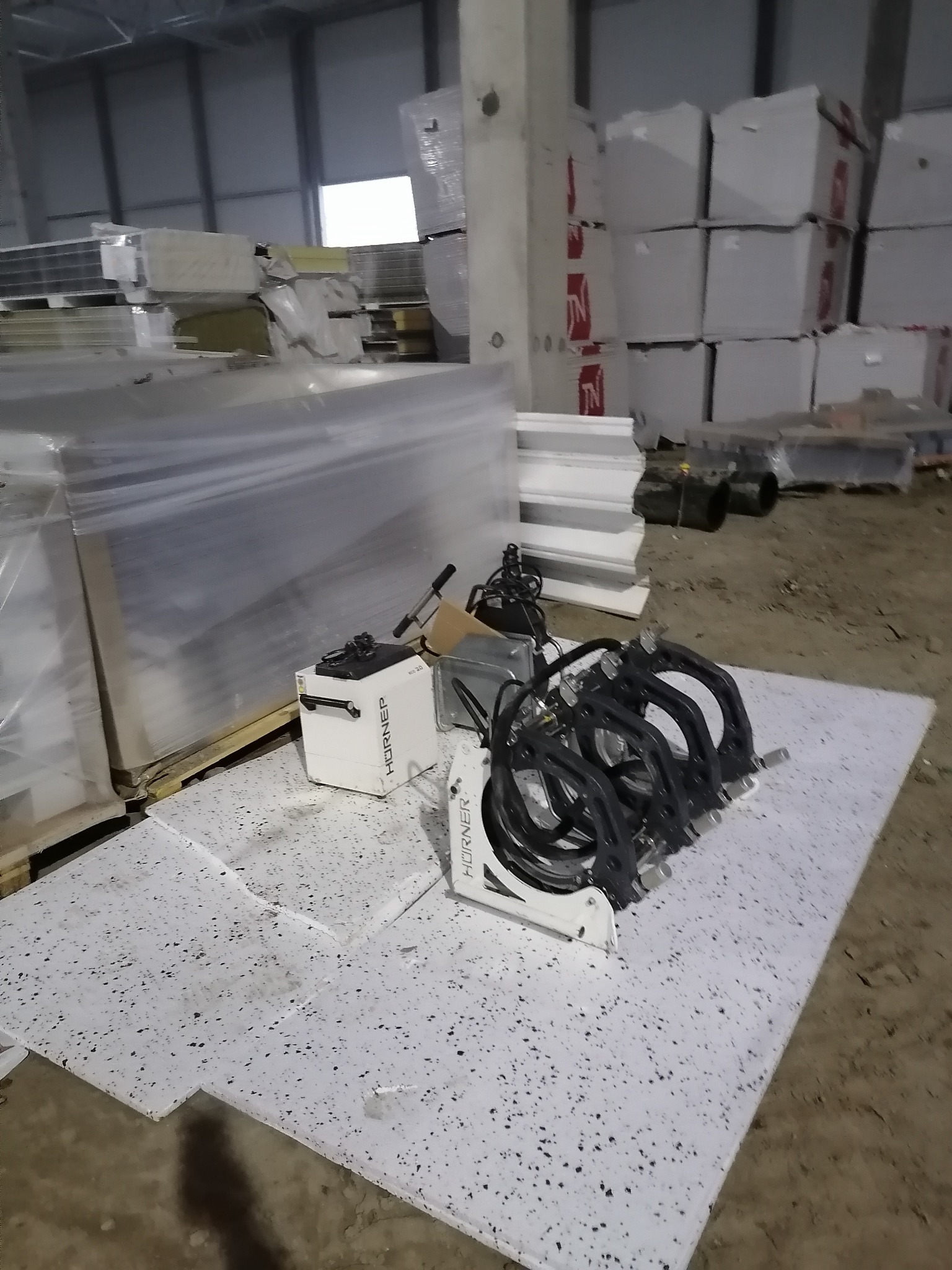 Magazynowe ujęcie: biała maszyna HÖRNER z czarnymi przewodami, obok palety zafoliowanych płyt, w tle stosy opakowań i betonowy słup.