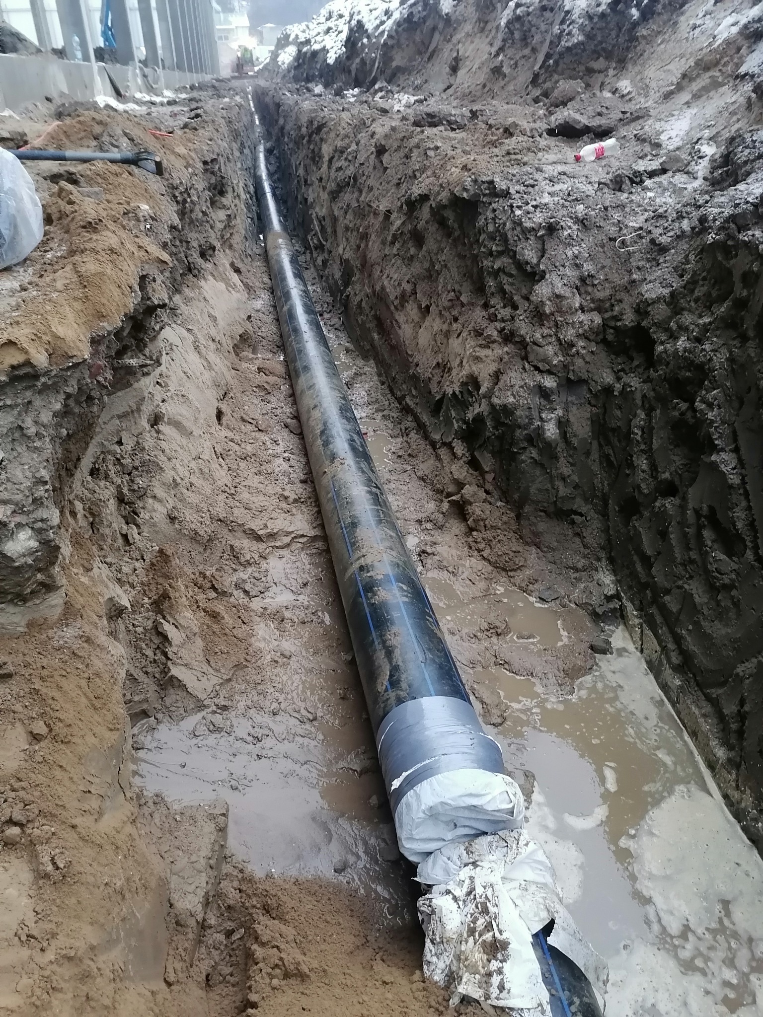 Długi, czarny rurociąg z owiniętymi końcami, ułożony w głębokim, wąskim wykopie w ziemi, z widocznymi śladami wody i błota w rowie.