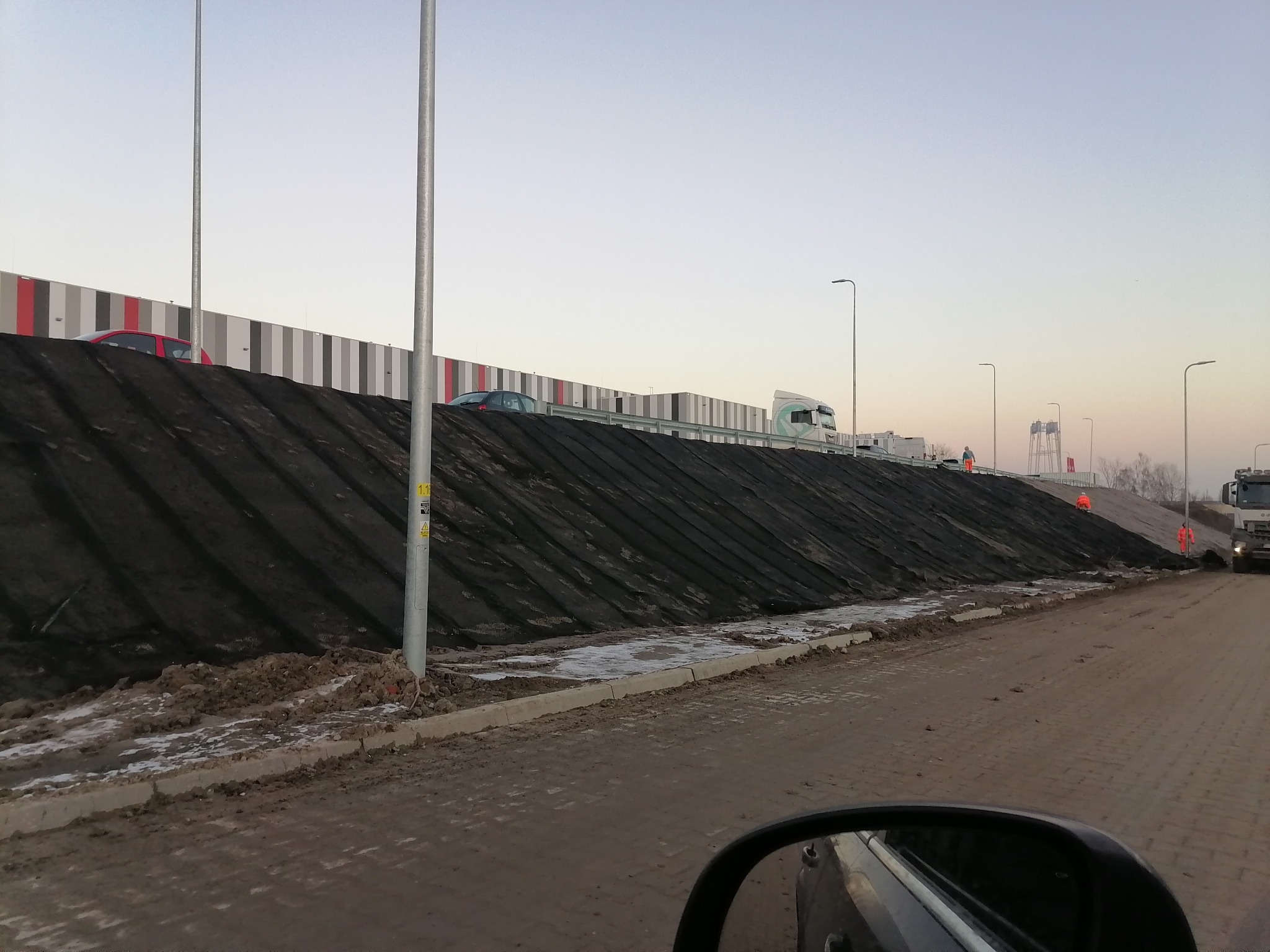 Skarpa zabezpieczona czarną geowłókniną, widoczni pracownicy w odblaskowych kombinezonach, w tle budynek przemysłowy z panelami w kolorach szarości i czerwieni, fragment samochodu w dolnym rogu kadru.