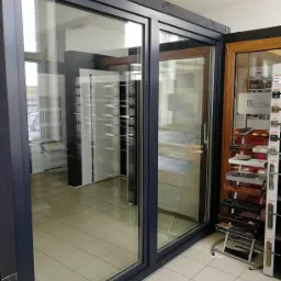 Wystawa okien z ciemnymi ramami i dużymi przeszkleniami, widoczne akcesoria i ekspozytory z klamkami.