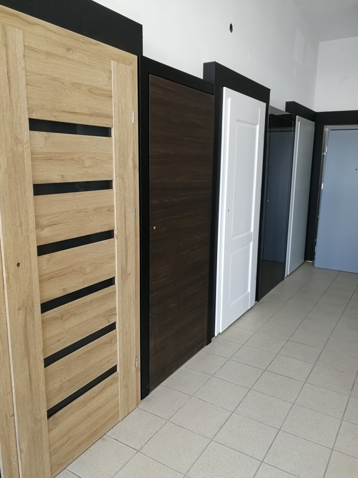 Ekspozycja różnych modeli drzwi wewnętrznych i zewnętrznych: drewnopodobne z czarnymi wstawkami, ciemnobrązowe, białe panelowe i szare, ustawione w rzędzie na tle podłogi z płytek.