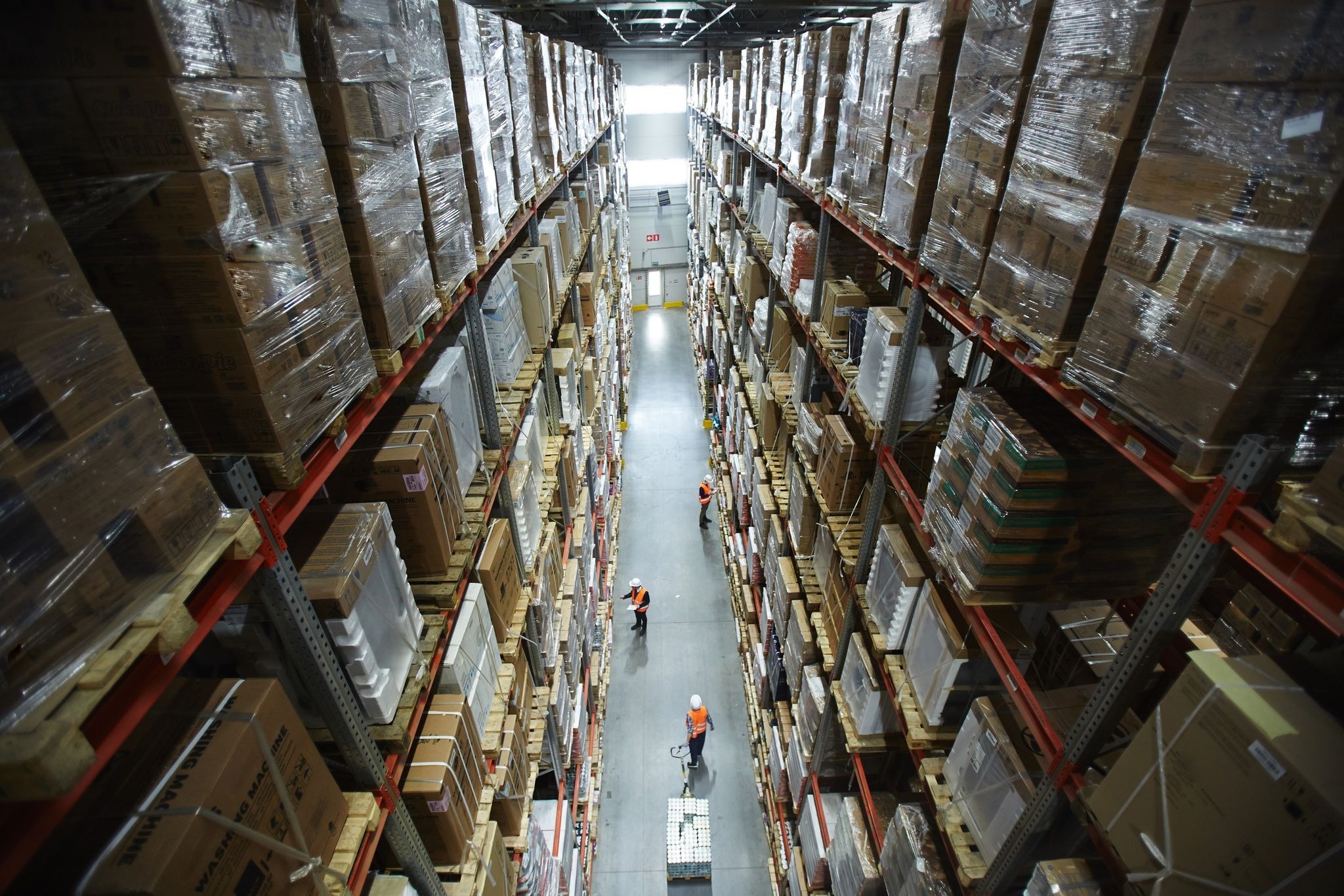 Wysoki magazyn z rzędami regałów wypełnionych kartonami i towarami owiniętymi folią, widok z góry na alejkę z pracownikami w odblaskowych kamizelkach i kaskach.
