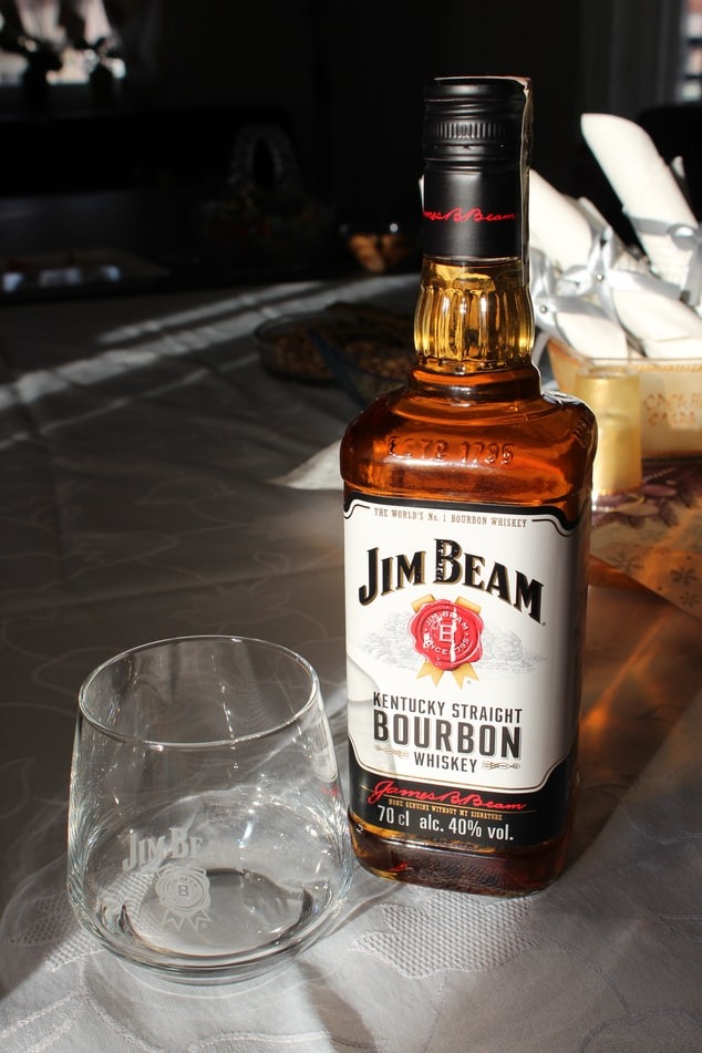 Butelka bourbonu Jim Beam obok pustej szklanki z logo firmy na stole w słoneczny dzień.