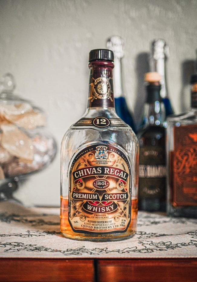 Butelka Chivas Regal Premium Scotch Whisky z numerem 12 na etykiecie, ustawiona na wzorzystej serwetce z innymi butelkami w tle, prezentowana w eleganckim otoczeniu.
