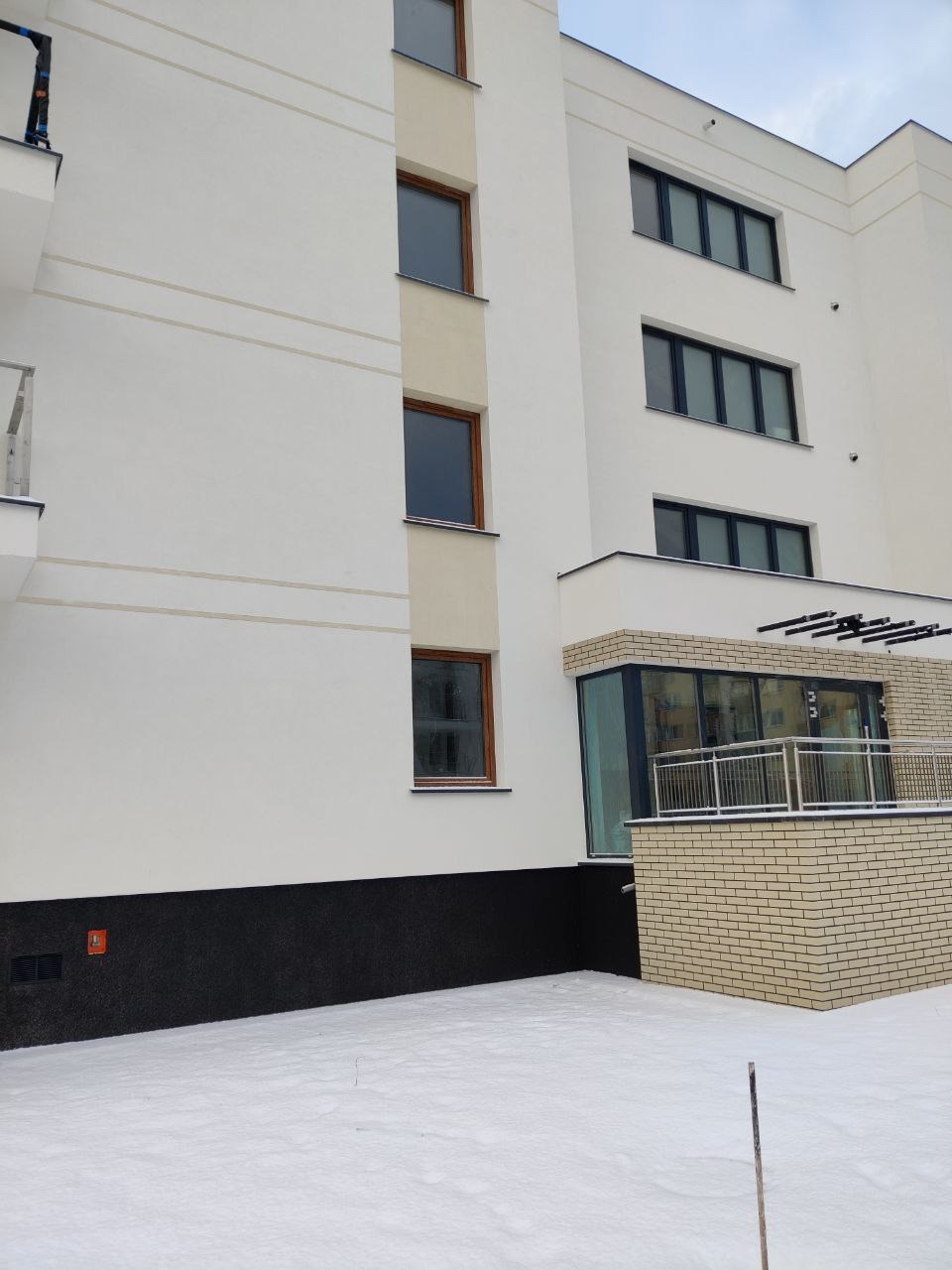 Nowoczesny budynek wielorodzinny z białą elewacją, ciemną podmurówką i kremowymi wstawkami, widoczny w zimowej scenerii z pokrywą śnieżną.