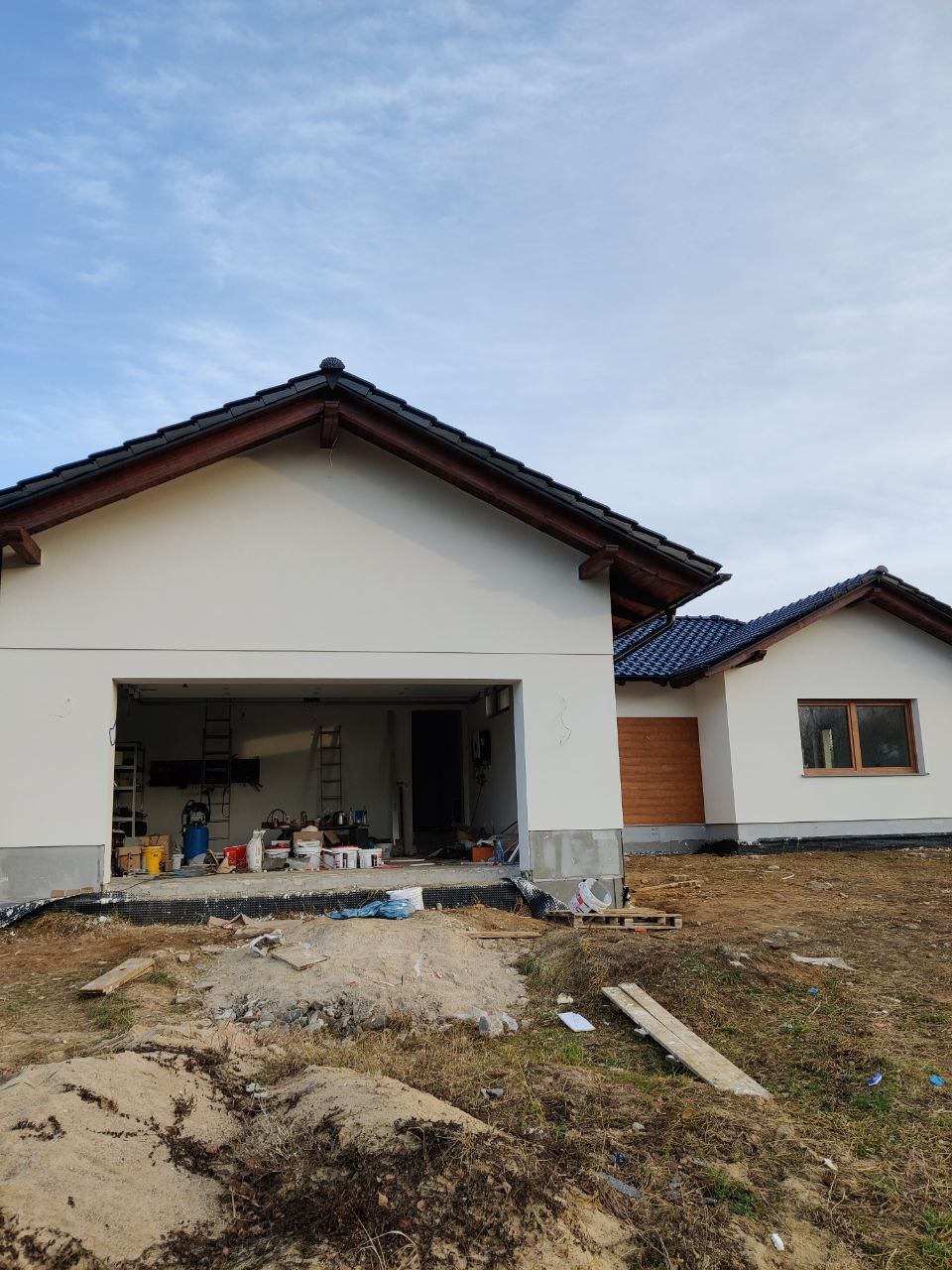 Niedokończony dom murowany z otwartym garażem, widoczne materiały budowlane i drabiny wewnątrz, teren wokół w trakcie prac ziemnych.