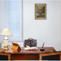 Sala konferencyjna z drewnianym biurkiem, otwartą książką, tabletem w etui, piórem w stojaku i skórzaną aktówką na tle białej ściany z obrazem i regałem z książkami.