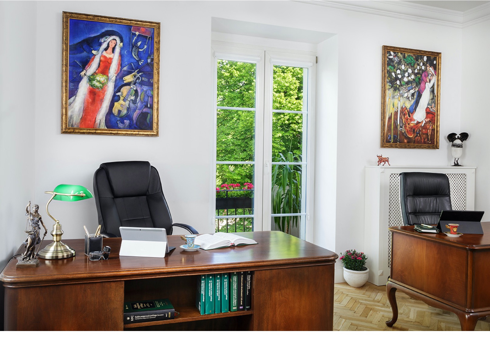 Jasne biuro z drewnianym biurkiem, otwartą książką, tabletem, lampką z zielonym kloszem i figurką Temidy; na ścianach wiszą obrazy w złotych ramach, a za oknem widać zieleń drzew.
