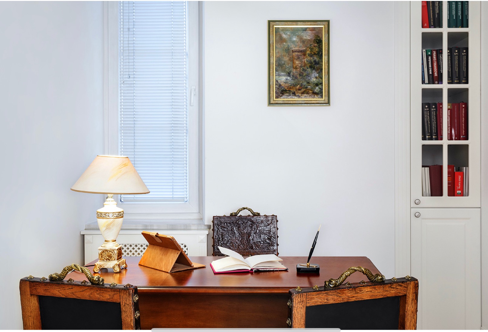Sala konferencyjna z drewnianym biurkiem, otwartą książką, tabletem w etui, piórem w stojaku i skórzaną aktówką na tle białej ściany z obrazem i regałem z książkami.