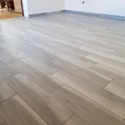 Ułożona podłoga z płytek imitujących drewno w odcieniach szarości, z szarą listwą przypodłogową, widok z perspektywy podłogi w kierunku otwartych drzwi do kolejnego pomieszczenia.