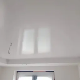 Biały, świeżo pomalowany sufit z delikatnym połyskiem, widoczne odbicie światła z okna, hak wystający z sufitu, narożnik pokoju z oknem.