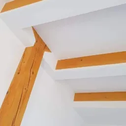 Biały sufit z drewnianymi belkami i skośną podporą, widoczne połączenie belek ze ścianą i sufitem, minimalistyczny design.
