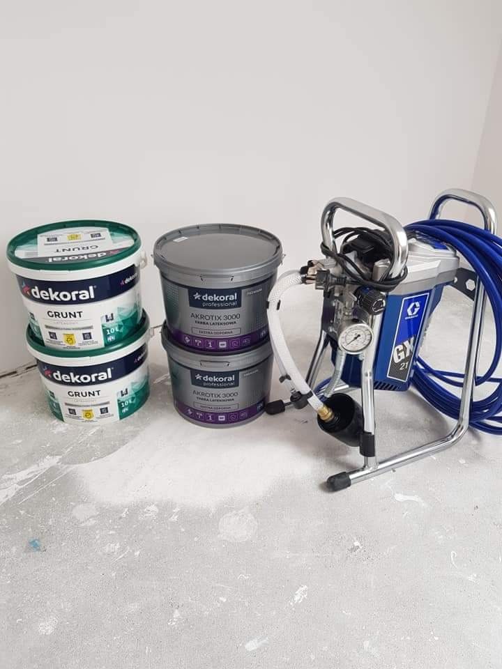 Agregat malarski Graco GX21, dwa wiadra farby lateksowej Dekoral Akrotix 3000 i dwa wiadra gruntu Dekoral ustawione na podłodze z widocznymi śladami farby.