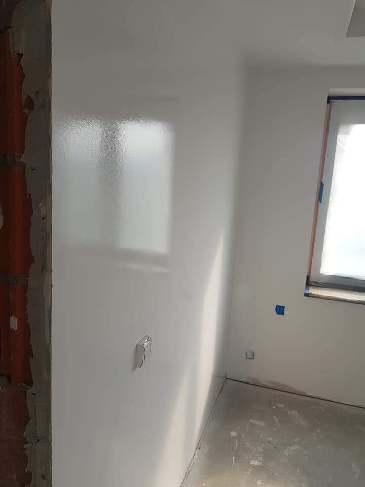 Świeżo pomalowane na biało wnętrze pokoju z widoczną ścianą z cegły w rogu, zabezpieczonym oknem i szarym betonowym podłożem, w trakcie prac wykończeniowych.