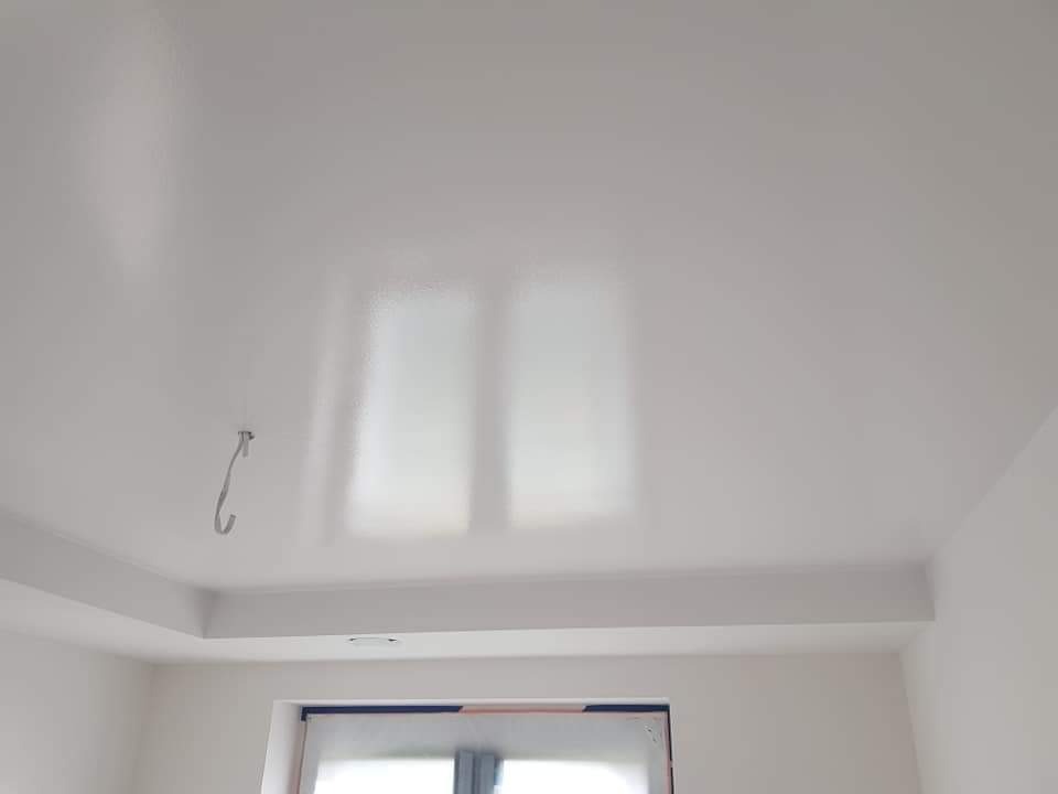 Biały, świeżo pomalowany sufit z delikatnym połyskiem, widoczne odbicie światła z okna, hak wystający z sufitu, narożnik pokoju z oknem.