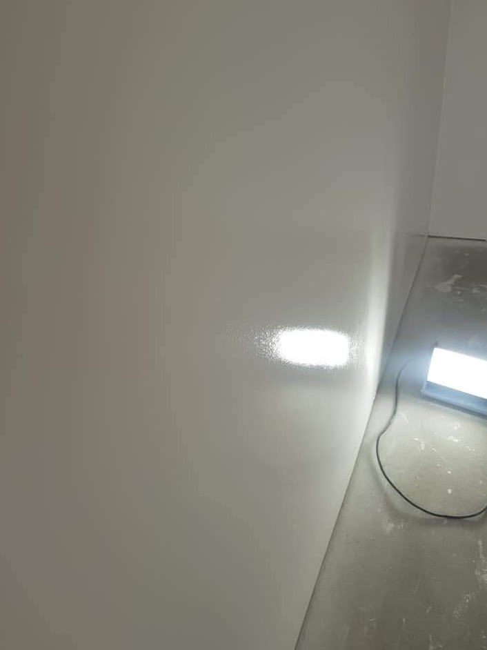 Świeżo pomalowana ściana w odcieniach szarości, oświetlona lampą budowlaną, widoczny fragment podłogi w trakcie prac wykończeniowych.