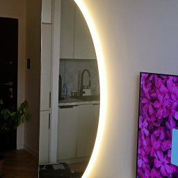 C.K.Glass - Okrągłe lustro z podświetleniem LED w kształcie półksiężyca, odbijające wnętrze kuchni i fragment telewizora z motywem kwiatowym.