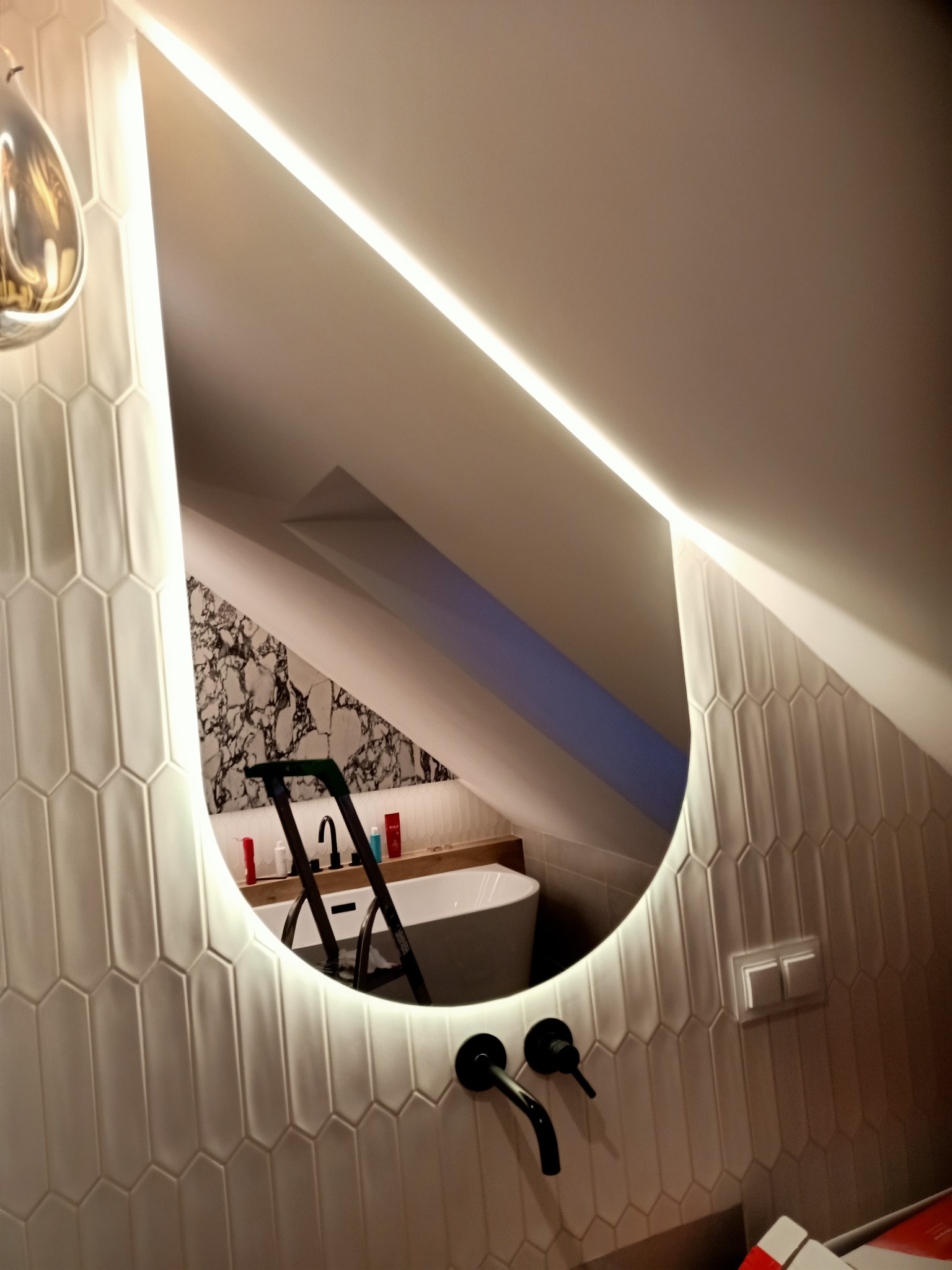 Oświetlone lustro w kształcie łzy odbija remontowane wnętrze łazienki w Gdańsku, widoczna drabina, wanna i marmurowe płytki na ścianie pod skosem.