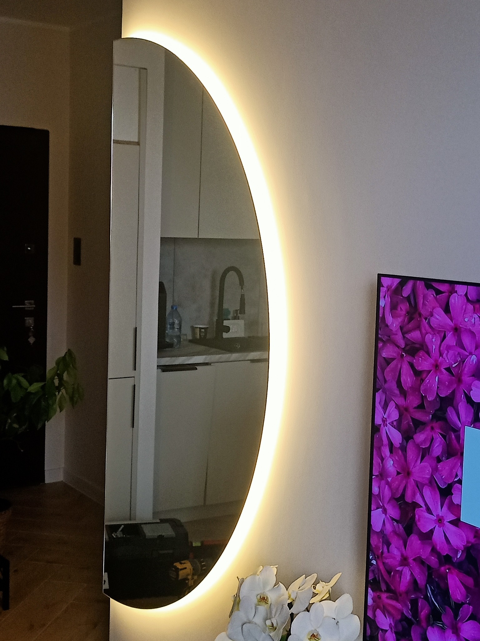 Okrągłe lustro z podświetleniem LED w kształcie półksiężyca, odbijające wnętrze kuchni i fragment telewizora z motywem kwiatowym.
