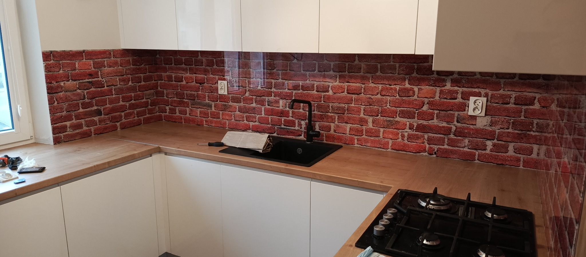 Szklany panel kuchenny z motywem cegieł nad blatem i zlewem, widoczne gniazdka elektryczne, czarny kran i fragment płyty gazowej.