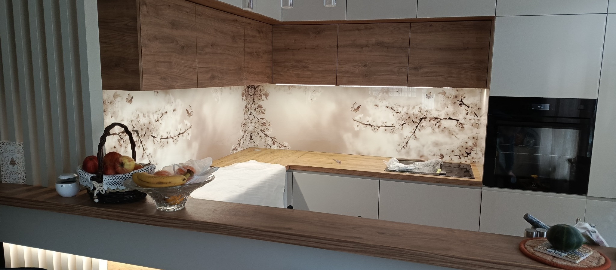 Szklana ściana w kuchni z motywem kwitnącej wiśni i motyli, brązowe szafki wiszące, blat z jasnego drewna, owoce w koszyku na pierwszym planie.